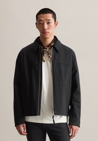 Schwarze Zip-Jacke mit weicher Textur, ausgestattet mit einem kragenförmigen Halsausschnitt und seitlichen Taschen, getragen über einem weißen Hemd und kombiniert mit einem gemusterten Schal.