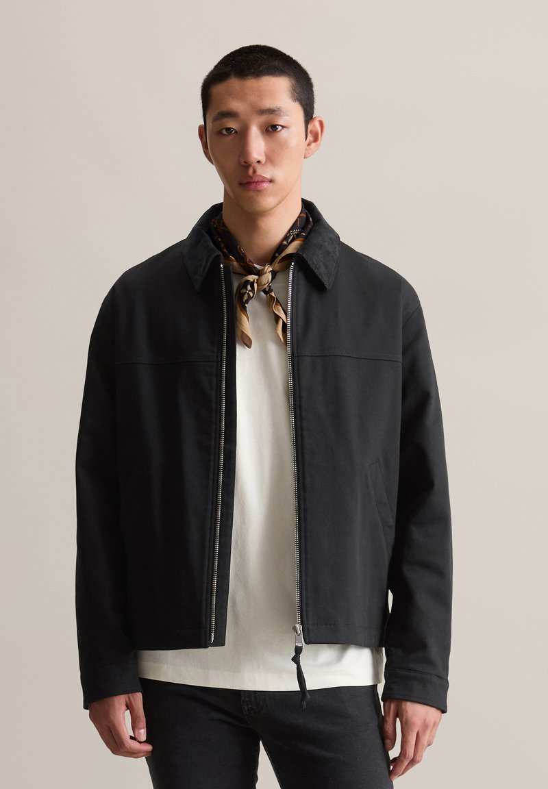 Schwarze Zip-Jacke mit weicher Textur, ausgestattet mit einem kragenförmigen Halsausschnitt und seitlichen Taschen, getragen über einem weißen Hemd und kombiniert mit einem gemusterten Schal.