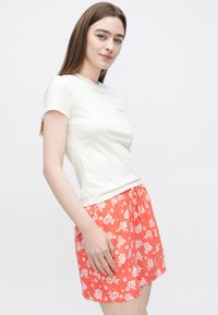 Bílé bavlněné tričko s krátkými rukávy a malým logem, v kombinaci s červenými šortkami s bílým květinovým vzorem a elastickým pasem.