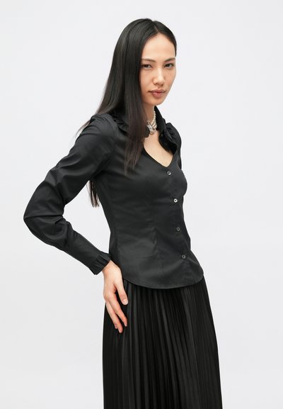 Melna pieguļoša krekls ar volānveida apkakli, gariem piedurknēm un pogu aizdari. Kombinēta ar melnu plisuētu svārku. Gluda tekstūra.