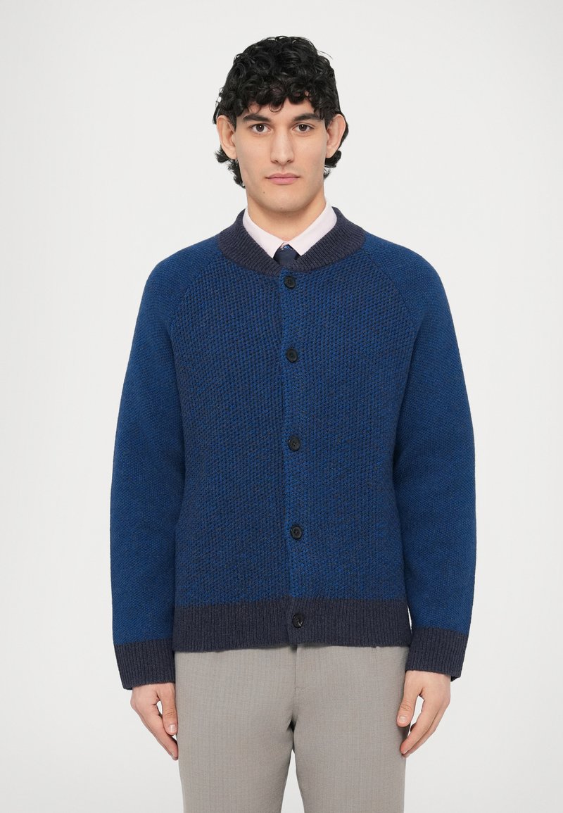 Dunkelblauer Pullover mit strukturiertem Strickmuster, Raglanärmeln und einem dunklen Kragen. Fünf schwarze Knöpfe vorne entlang.