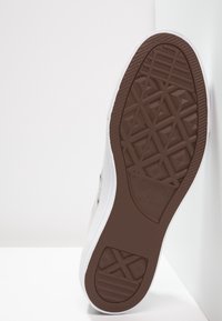 Brun gummisula med texturerat geometriskt mönster, vit kant och logotyp. Ljust grå överdel med subtil textur och rundad tådesign.