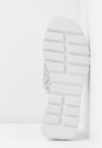 Semelle en caoutchouc blanc avec des rainures profondes et un motif texturé, tige de couleur claire partiellement visible avec un design en peau de reptile, sur un fond blanc uni.