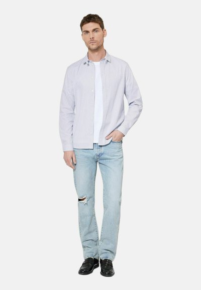 Homme portant une chemise grise clair boutonnée sur un t-shirt blanc, un jean bleu clair déchiré et des chaussures noires, debout avec une main dans la poche.