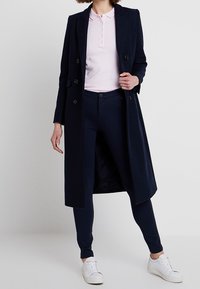 Marineblå lang frakke med dobbeltknappede knapper, båret over en lyserød polo og mørke bukser, parret med hvide sneakers. Glat stof, skræddersyet pasform.