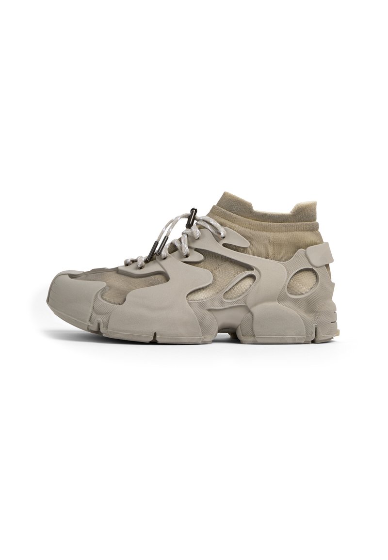 Scarpa sportiva beige con tomaia in gomma texturizzata e rete, colletto elastico e design con ritagli distintivi. Presenta lacci grigi e accenti di supporto.