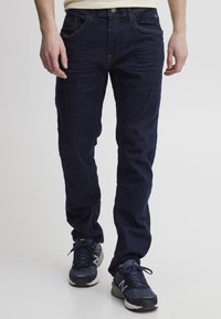 Jeans en azul oscuro con un diseño ajustado y un ligero efecto de desgaste, complementados con costuras en naranja y un botón plateado. Combinados con zapatillas deportivas oscuras.