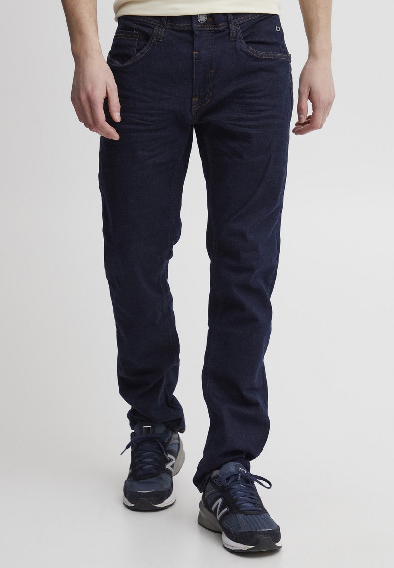 Jeans en azul oscuro con un diseño ajustado y un ligero efecto de desgaste, complementados con costuras en naranja y un botón plateado. Combinados con zapatillas deportivas oscuras.