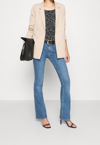 Beige jakke over en marineblå top med bladmønster, blå ispanske jeans, sort bæltet, og sorte strop sandaler. Minimalistisk sort håndtaske.