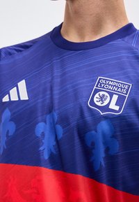Μπλε και κόκκινη φανέλα ποδοσφαίρου Olympique Lyonnais με το έμβλημα της ομάδας και το λογότυπο της Adidas στο στήθος, φορεμένη από ένα άτομο που φαίνεται από τον λαιμό έως τη μέση του κορμού.