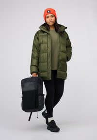 Giacca a vento verde isolata con cappuccio, abbinata a un maglione verde scuro, leggings neri e uno zaino nero. Berretto arancione. Sneakers nere.