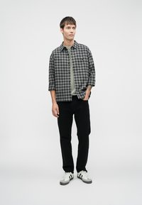 Camisa de cuadros gris y negra de manga corta, con cierre de botones, usada sobre una camiseta verde; combinada con pantalones negros y zapatillas blancas.