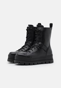 Bottes en cuir noir à la cheville avec une semelle plateforme épaisse, lacets à l'avant et une sangle logo sur le côté. Surface texturée et languette de tirage incluses.
