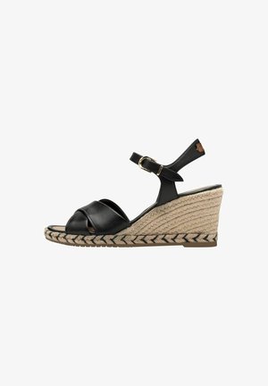 Black leather wedge sandal with crossover straps, textured jute platform and ankle strap featuring a gold buckle, accented by a black pattern.