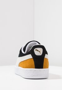 Svart mockaskor med orange och vita accenter. Har en texturerad vit sula och en guldfärgad Puma-logotyp på hälen. Klassisk låg sko-design.