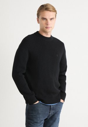 Maglione nero a costine con scollatura rotonda e maniche lunghe, caratterizzato da un piccolo logo ricamato sul petto, abbinato a jeans di denim blu.