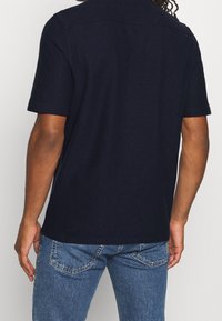 Polo de manga corta en color navy con tejido texturizado, cuello estándar y dobladillo recto. Combinado con jeans de mezclilla en tono azul claro.