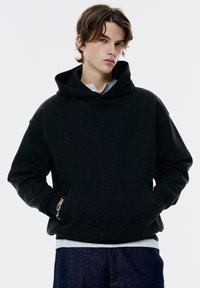 Giovane con capelli castani, vestito con un felpa nera e jeans blu, in piedi con le mani nelle tasche della felpa su uno sfondo semplice.