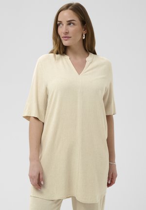 Kobieta o jasnobrązowych włosach, ubrana w beżową luźną tunikę z rękawami do łokcia i dekoltem w kształcie litery V, patrząca lekko w lewo.