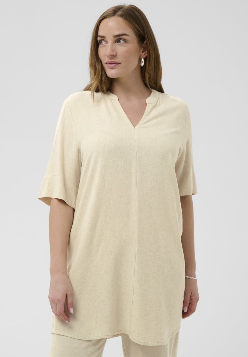 Világosbarna hajú nő, aki bézs színű, laza tunikát visel, melynek könyékig érő ujjai és V-alakú kivágása van, kissé balra néz.