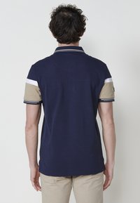 Tengerészeti póló, bézs és fehér csíkos ujjakkal, két gombos gallérral és strukturált anyaggal. Klasszikus fazon, oldalsó hasítékokkal.