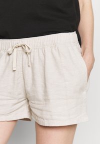 Pantalones cortos de un beige claro con cintura elástica y cordón. Elaborados en un tejido texturizado con patrones de rayas verticales. Bolsillos laterales visibles.