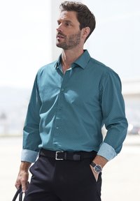 Türkises Hemd mit Knopfleiste und einem kleinen weißen Punktmuster; kontrastierende hellblaue Manschetten; kombiniert mit einem schwarzen Gürtel und dunklen Hosen.