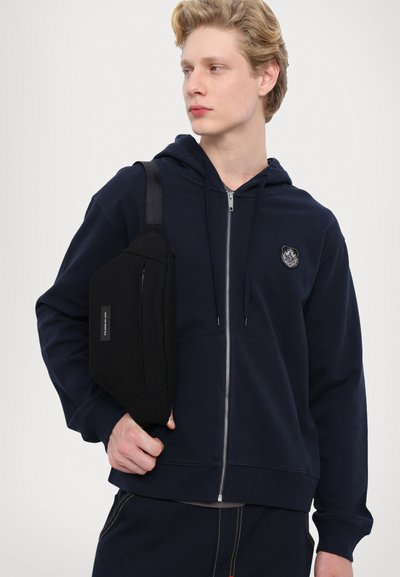 Tumesinane kapuutsiga navy värvi jakk hõbedase tõmblukuga, millel on ees embleem ja ribikud varrukatel. Must rihmakott, millel on tõmblukuga tasku, kantakse kehale ümber.