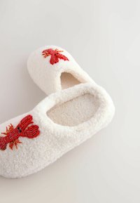 Weiße, flauschige Hausschuhe mit offener Rückseite, die vorne mit roten, perlenbesetzten Hummer-Motiven verziert sind. Weicher Stoff mit runder Form.