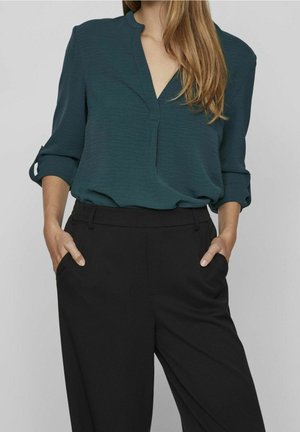 Kobieta w zielono-niebieskiej bluzce z dekoltem w serek i podwiniętymi rękawami, włożonej do wysokich czarnych spodni, z rękami w kieszeniach.