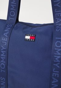 Bolso de mano azul marino con cierre de cremallera, que presenta una tela texturizada y un pequeño parche con el logo. Las asas exhiben "TOMMY JEANS" en relieve.