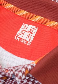 Rode en gestippelde stof met oranje accenten bevat een label met "J&JOY," een Union Jack-logo en de tekst "UK Exploration."