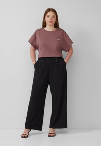 Weinrotes kurzärmliges T-Shirt mit einem kleinen Grafikmotiv, kombiniert mit hochgeschnittenen schwarzen weitem Hose mit vorderen Knöpfen.