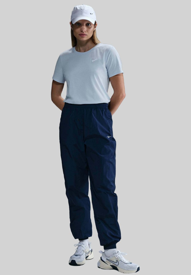T-shirt azzurro con logo, pantaloni con cintura elastica blu navy, cappellino bianco e sneakers bianche con dettagli neri. Abbigliamento sportivo casual.