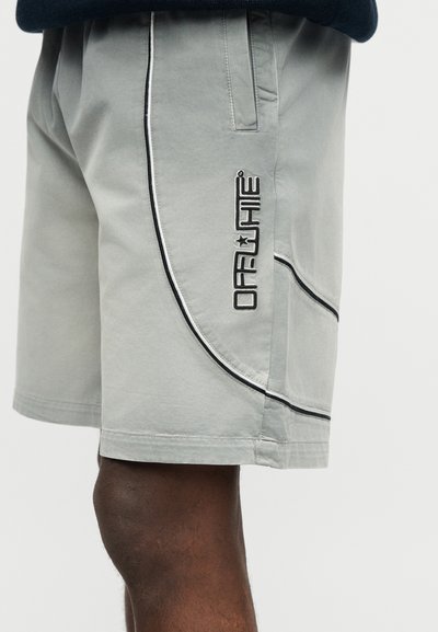 Ανοιχτό γκρι σορτς με μαύρες λεπτομέρειες και κάθετη ετικέτα "OFF-WHITE" στο πλάι, φορεμένα από ένα άτομο με σκούρο δέρμα.