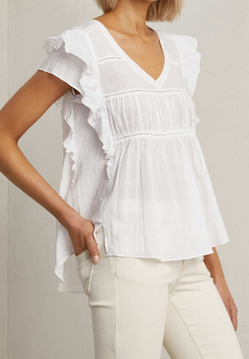 Vit, lätt blus med volangärmar, V-ringning, texturerad tyg och samlad detaljering. Matchas med ljusa, åtsittande byxor.