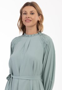 Rochie verde mentă cu o gâtă înaltă, strânsă, față pliată, mâneci lungi și un brâu din material la talie. Textură netedă.