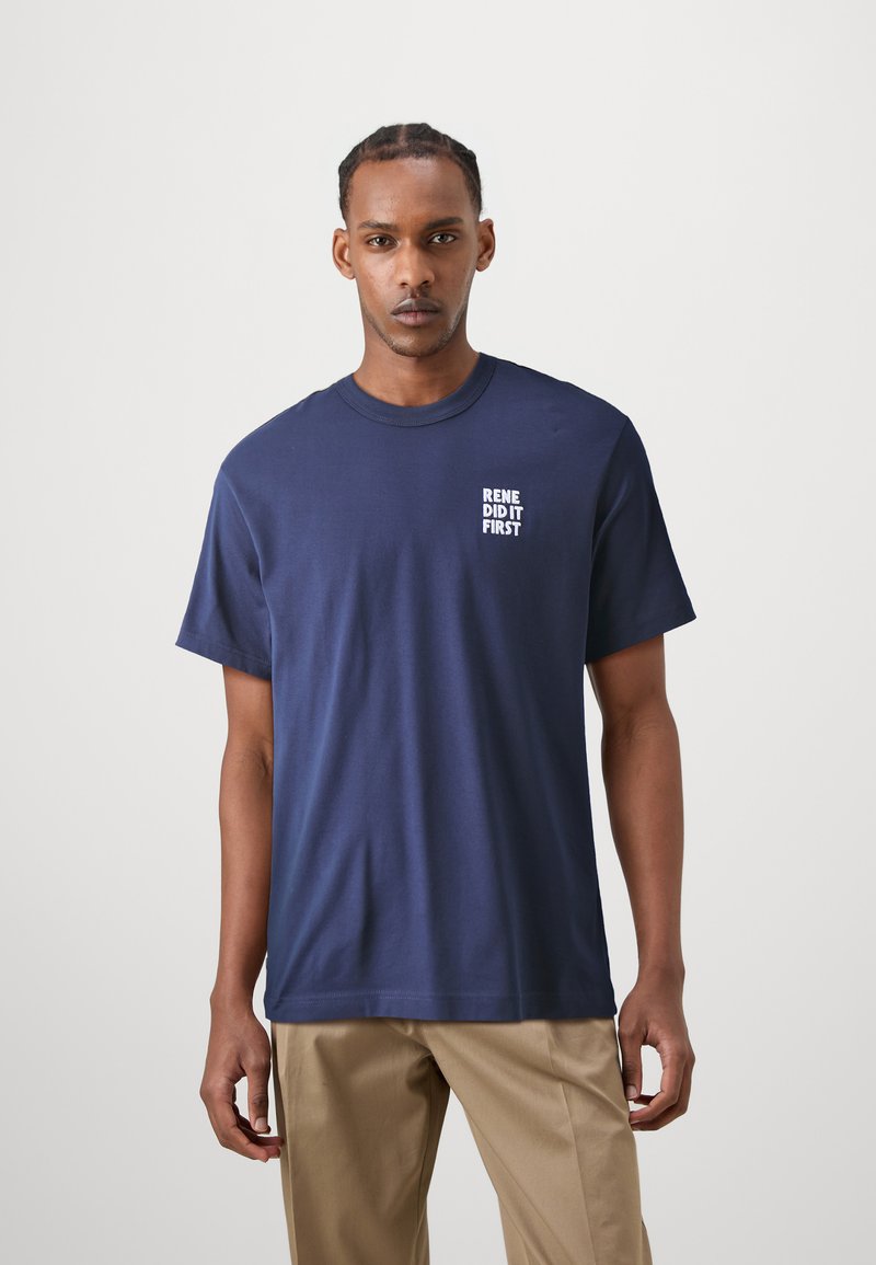 Camiseta de algodón azul marino con mangas cortas y cuello redondo; texto blanco "RENE LO HIZO PRIMERO" impreso en la parte izquierda del pecho.
