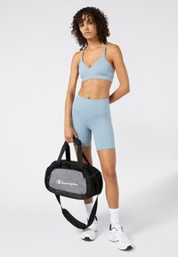 Sujetador deportivo azul claro y pantalones cortos con una textura suave. Bolsa de lona Champion en negro y gris con logotipo blanco. calcetines cortos blancos y zapatillas deportivas.