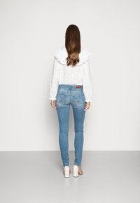 Blouse blanche texturée à col à volants, associée à un jean bleu clair ajusté et des chaussures blanches à talons hauts, vue de dos.