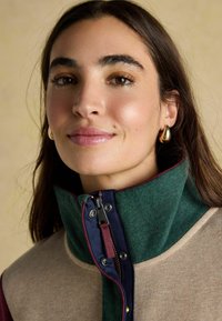 Mladá žena s hnedými vlasmi má na sebe béžovo-zeleno-námorníkový kabát s vysokým golierom a zlatými kruhovými náušnicami, jemne sa usmieva na neutrálnom pozadí.