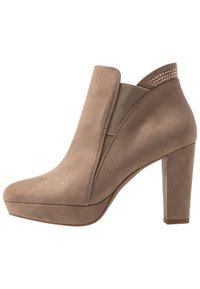 Beige mokkasini nilkkuri, jossa on korkea lohkoiltapohja, joustavat sivupaneelit ja koristeellinen niittikoristus takana. Sileä pinta, pyöristetty kärki.