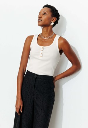 Femme portant un haut blanc sans manches avec des boutons, un pantalon large noir à motifs, des colliers superposés et des boucles d'oreilles en forme d'étoile, posant contre un mur blanc.