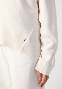 Light beige sweatshirt with a crew neckline, ribbed cuffs, and side detail. Soft texture and minimalistic design. Paired with white pants.
