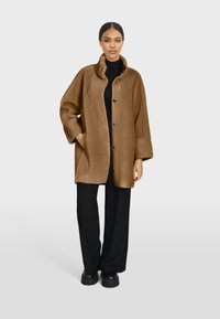 Manteau en fausse fourrure brune avec un col, des boutons à l'avant et des poches latérales. Coupe ample avec des manches larges, porté avec un pantalon noir et des chaussures épaisses.