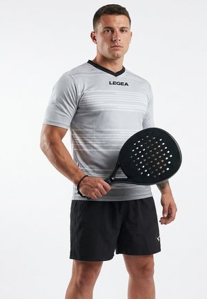 Junger Mann trägt graues Sportshirt und schwarze Shorts, hält einen schwarzen Padel-Schläger und steht vor weißem Hintergrund.