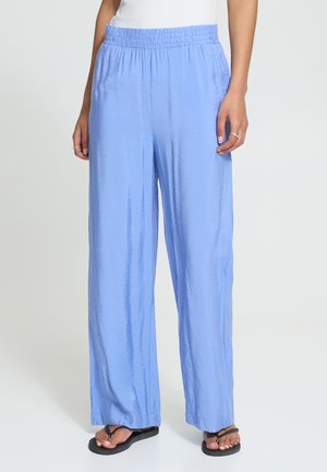 Frau trägt lockere, hellblaue Satinhose mit elastischem Bund und schwarzen Flip-Flops, steht vor weißem Hintergrund.