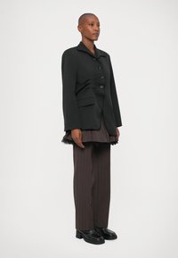 Blazer negro a medida con cuello de muesca, diseño de un solo botón y cuatro botones. Combinado con pantalones marrones con pliegues y zapatos negros.