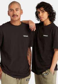 To personer iført svarte bomull t-skjorter med en liten hvit Timberland-logo. Personen til venstre har tatoveringer og bærer sølv smykker.