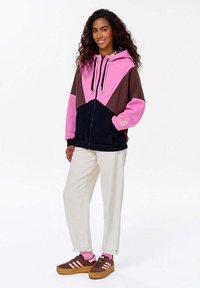Hoodie w kolorze blokowym w odcieniach różu, brązu i granatu. Posiada zamek błyskawiczny, dwie kieszenie oraz kaptur ze sznurkiem. Połączony z kremowymi spodniami i brązowymi trampkami.
